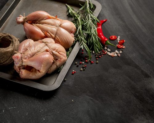 Young chicken on a baking sheet with seasonal vegetables and greens before baking in the oven. Photo of food on a dark background for a book of recipes or design. Horizontal view.